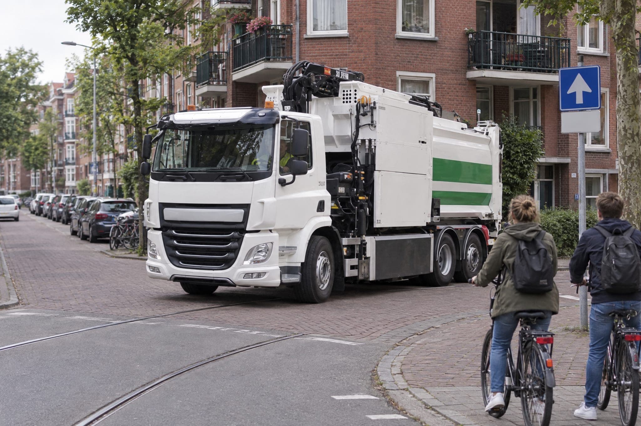 Refuse truck with a crane arm turning into a residential street while two cyclists wait at the corner